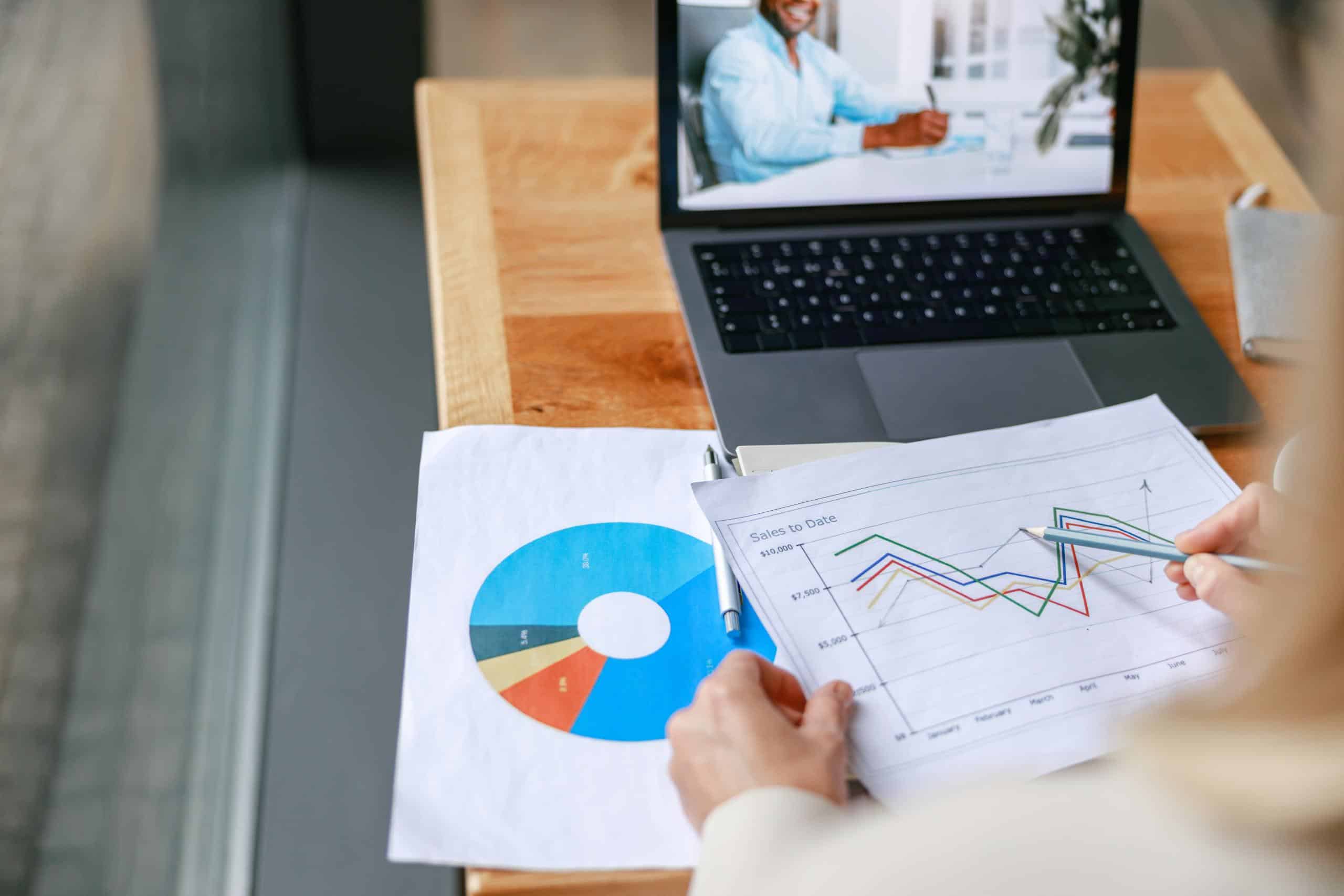 Analyse von Finanzdaten und Rechnungsprozessen: Geschäftsperson betrachtet Diagramme vor Laptop im Video-Meeting zur Digitalisierung der Buchhaltung.